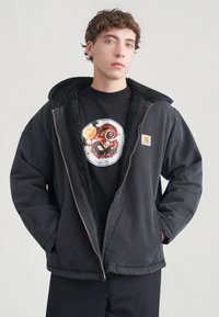 Μαύρο μπουφάν puffer με υφασμάτινη κουκούλα επενδυμένη με φλις. Φοριέται πάνω από ένα μαύρο μπλουζάκι με πολύχρωμο κυκλικό γραφικό και κλείσιμο με φερμουάρ.