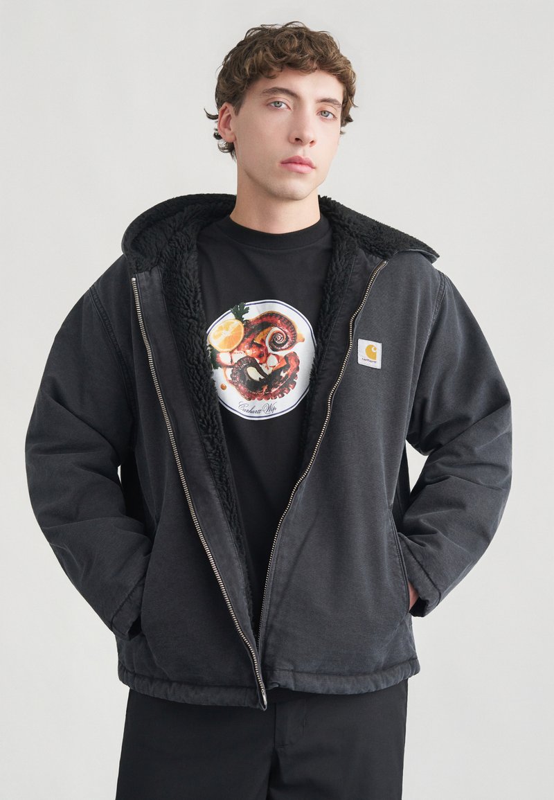 Sort pufferjakke med hætte foret med tekstureret fleece. Bæres over en sort t-shirt med en farverig cirkulær grafik og lynlåslukning.