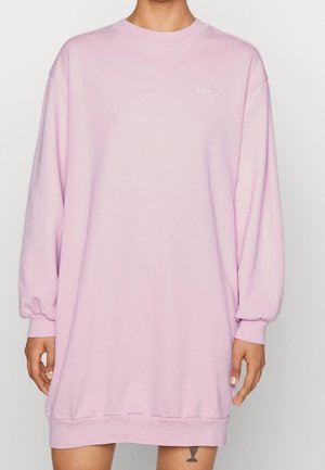 Personne portant une robe-sweat-shirt rose pâle à manches longues oversized avec un petit logo blanc Levi's sur la poitrine, debout devant un fond uni.