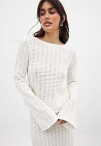 Valge kudumine särk, millel on lahti ja mugav siluett, laiad kellukesed varrukad ja tekstureeritud avatud kudumise muster.