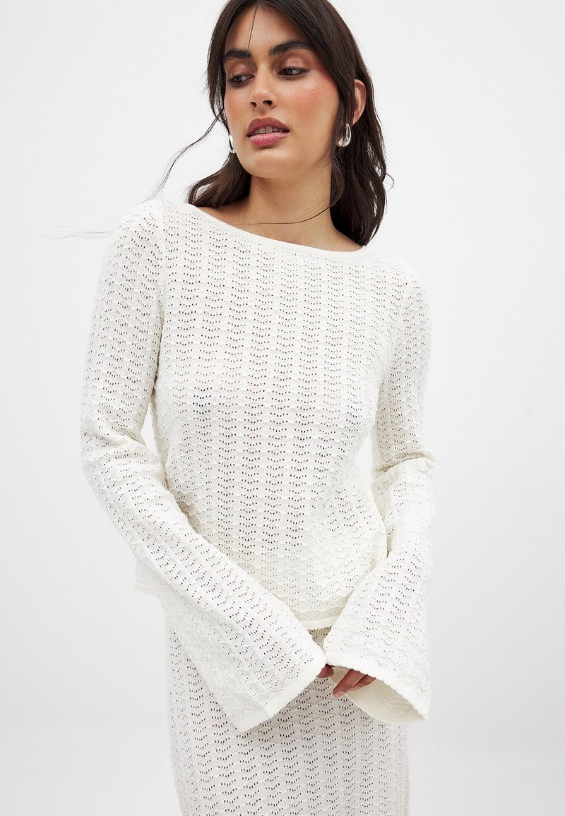 Valge kudumine särk, millel on lahti ja mugav siluett, laiad kellukesed varrukad ja tekstureeritud avatud kudumise muster.