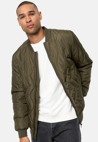 Chaqueta bomber acolchada en verde oliva, con cierre de cremallera en frente, puños acanalados y relleno ligero. Llevada sobre una camisa blanca.