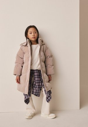 Junges Mädchen trägt eine beige Steppjacke, ein weißes Hemd, weiße Hose und ein gebundenes lila kariertes Hemd und steht vor einer einfarbigen beigen Wand.