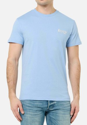 Mann trägt hellblaues kurzärmeliges T-Shirt mit weißem "Boxeur Des Rues"-Logo auf der Brust und mittelblauen Jeans.