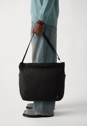 Black fabric messenger bag with a smooth texture, adjustable shoulder strap, and zippered closure, held by a person wearing light blue clothing.