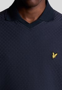 Marinblå polo skjorta med en texturerad mönster, med en V-ringning och en gul broderad logotyp på vänster bröst. Mjuk kvalitet.