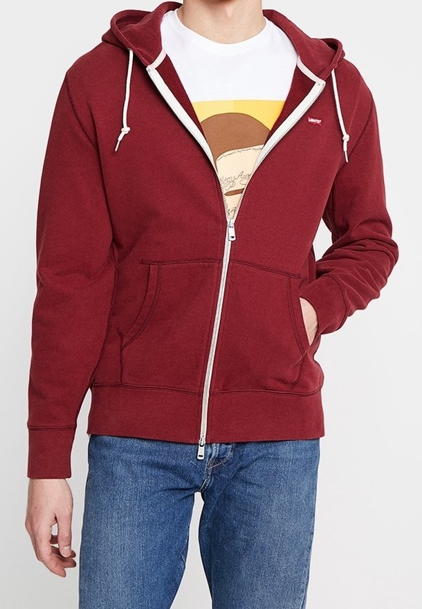 Personne portant un sweat à capuche bordeaux zippé avec des cordons blancs sur un t-shirt graphique blanc et un jean bleu, main dans la poche du sweat.