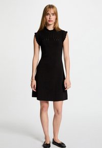 Black sleeveless dress with a fitted bodice, scalloped neckline, and flared hem. Smooth texture and minimalist design. Paired with black flat shoes.