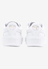 Bílé kožené tenisky s texturovaným povrchem, která mají vyvýšenou bílou podrážku a zlaté logo na zadní straně a na jazyku.