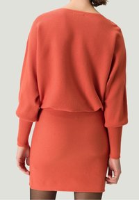 Robe en tricot côtelé corail avec un design à épaules tombantes. Manches longues et jupe ajustée, mettant en valeur une surface texturée.