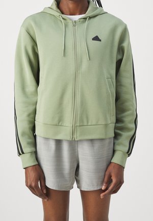 Person iført en lysegrøn hættetrøje med lynlås med sorte striber på ærmerne og lille logo, parret med lysegrå træningsshorts.