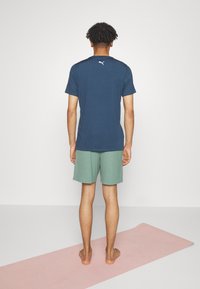 Námořnická modrá sportovní tričko s krátkým rukávem, spárované se zelenými žebrovanými šortkami. Stojí na růžové jógové podložce, viditelné bosé nohy.