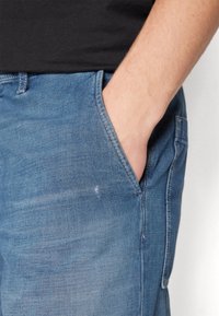 Högerhand vilande i framfickan på ljusblå denimjeans, bärande en svart skjorta.