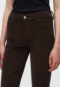 Gros plan d'une personne portant un pantalon en velours côtelé marron foncé avec des poches avant et un bouton argenté, associé à un haut noir sans manches.