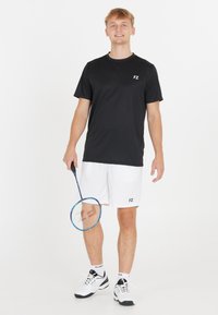 Svart idrottströja med FZ-logga, vita shorts med FZ-logga, håller en blå badmintonracket och har på sig svarta och vita skor.