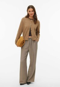 Brązowy dziany kardigan z złotymi guzikami, w połączeniu z szarymi, szerokimi spodniami z kieszeniami po bokach. Pod jednym ramieniem trzymana jest torebka w kolorze beżowym.