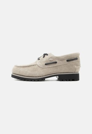 Chaussure bateau en daim beige avec lacets noirs, semelle noire à rainures profondes et œillets latéraux pour le détail des lacets.