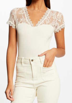 Kobieta ubrana w biały koronkowy top z krótkimi rękawami z falistymi brzegami oraz białe spodnie z wysokim stanem, ręka w kieszeni.