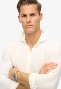Camisa blanca de lino con cuello, combinada con un reloj dorado y azul marino en la muñeca. El modelo tiene los brazos cruzados y un fondo neutro.