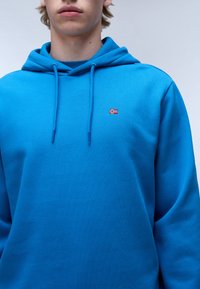Sudadera azul con una textura suave, cordones y un pequeño emblema de la bandera de Noruega en el pecho. Diseño sencillo con un corte relajado.