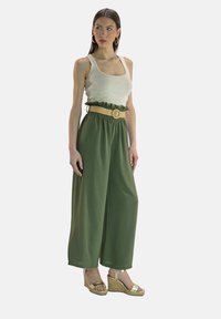 Gröna byxor med vida ben och volangkant vid midjan, kombinerade med en vit linne och beige stråbälte. Bärs med guldfärgade espadrillekilklackssandaler.