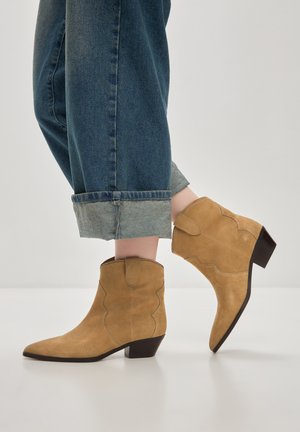 Botine din suede tan până la glezna, cu vârfuri ascuțite, design cu margini ondulate și toc bronzat. Asortate cu blugi închiși la culoare, cu manșete rulate.