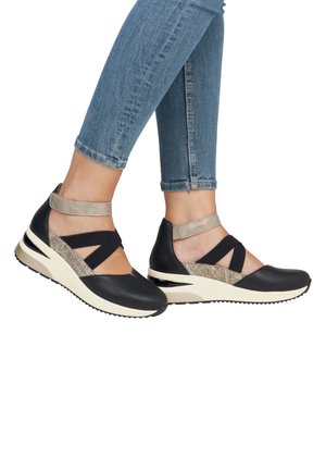 Jambes portant un jean bleu avec des chaussures compensées sportives noires et beiges, dotées de brides à la cheville et de détails texturés sur des semelles blanches.