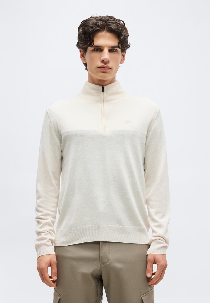 Mladý muž oblečený v svetlobéžovom pulóvri so štvrťzipsom a kaki nohaviciach stojaci pred jednoliatym bielym pozadím.