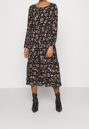 Femme portant une robe midi noire à manches longues avec un motif floral rouge, blanc et vert, associée à des bottines noires.