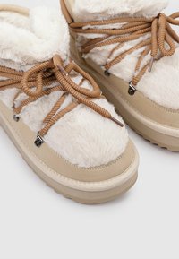 Zapatillas beige con paneles de crema esponjosos, con cordones marrones y detalles metálicos. La suela es gruesa y texturizada para proporcionar agarre.