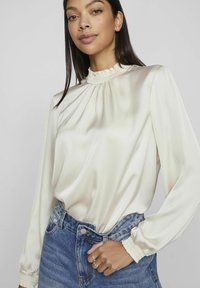 Silkebluse i off-white med en høj rynket krave og lange ærmer; parret med denimjeans. Glat tekstur, falder elegant.