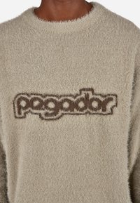 Világosbarna bolyhos pulóver kerek nyakkal, sötétbarna logóval hímezve a mellkason. Puha tapintású és laza fazonú.