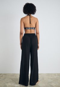 Crni bikini top bez naramenica sa brendom 'CALVIN KLEIN', u kombinaciji sa širokim crnim hlačama, koje imaju glatku teksturu i ležeran kroj.