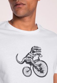 Bílá tričko s černým potiskem dinosaura na kole. Design se vyznačuje detailními linkami a hravým, kresleným stylem.