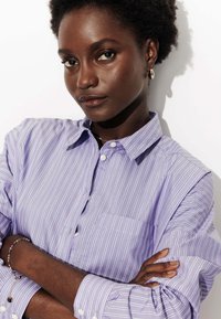 Chemise à rayures violettes claires, à boutons, à manches longues, avec une poche poitrine. Elle présente des rayures verticales blanches et un col.