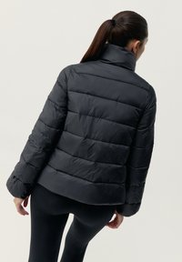 Črna puhovka z visokim ovratnikom, ki ima prešite dele in gladko teksturo. Zadnji pogled prikazuje sproščen kroj in dolge rokave.