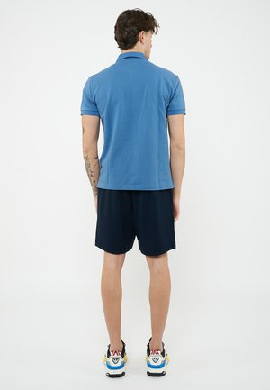 Uomo visto di spalle, indossa una polo blu, pantaloncini neri e sneakers multicolori, con tatuaggi sul braccio destro.
