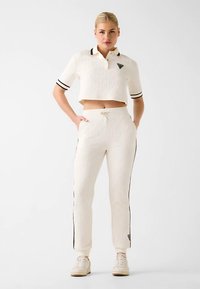 Cropped white polo with black stripes, logo on chest, paired with white pants featuring side stripes, drawstring waist, and textured fabric.