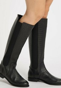 Cizme negre până la genunchi, cu un model texturat de crocodil, panouri elastice, toc plat, vârf rotunjit și fermoar pe lateral.