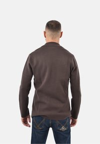 Uomo con capelli corti che indossa una giacca marrone a maniche lunghe e jeans blu, in piedi di spalle su uno sfondo semplice.