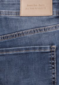 Primo piano della tasca posteriore di jeans in denim blu con etichetta in pelle beige con la scritta "street One - Denim ALL TIME FAVOURITE."
