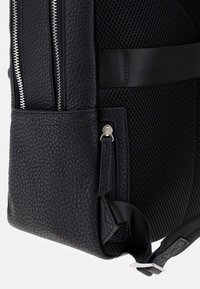 Zaino in pelle nera con superficie testurizzata, doppie cerniere, pannello in rete e spallacci regolabili. Presenta una tasca con cerniera e un design minimalista.