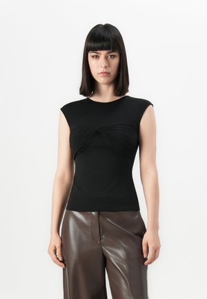 Woman with straight dark hair wearing a black sleeveless top with twist detail and shiny brown wide-leg pants against a white background.