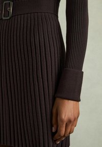 Stickad klänning i mörkbrunt med vertikal ribbning, midja med bälte samt långärmar med manschetter, vilket framhäver den texturerade tyget.