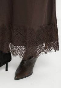 Brun lang kjole med intrikate blonde detaljer ved kanten og teksturert stoff. Svart støvlett med spiss tå synlig ved siden av.