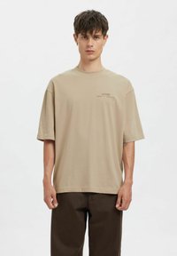 Béžové oversized tričko z měkké látky. Kulatý výstřih, krátké rukávy a malý potisk loga na levé straně. Rovný lem.