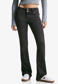 Black denim flared jeans with five pockets, metal buttons, and a mid-rise waist, paired with white sneakers and a gray cropped sweater.
