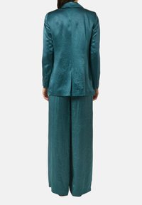 Kobieta ubrana w turkusowe, szerokie spodnie z teksturą i dopasowaną marynarkę, pokazana od tyłu na jednolitym białym tle.