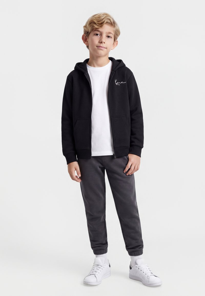 Gutt står iført svart hettejakke med glidelås, hvit t-skjorte, mørkegrå bukser, hvite sokker og hvite joggesko mot ensfarget bakgrunn.