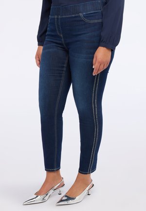 Frau in dunkelblauen Skinny-Jeans mit Seitennahtdetails und silbernen, spitzen Slingback-Highheels, stehend vor einem weißen Hintergrund.
